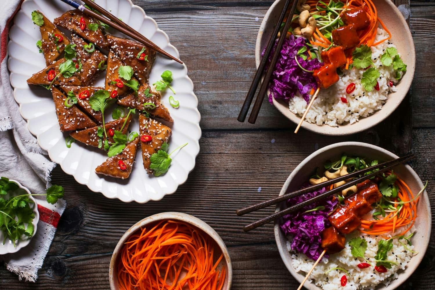 Vegetarian meal featuring triangular tofu pieces garnished with sesame seeds, cilantro, green onions, and red chili slices on a white plate.