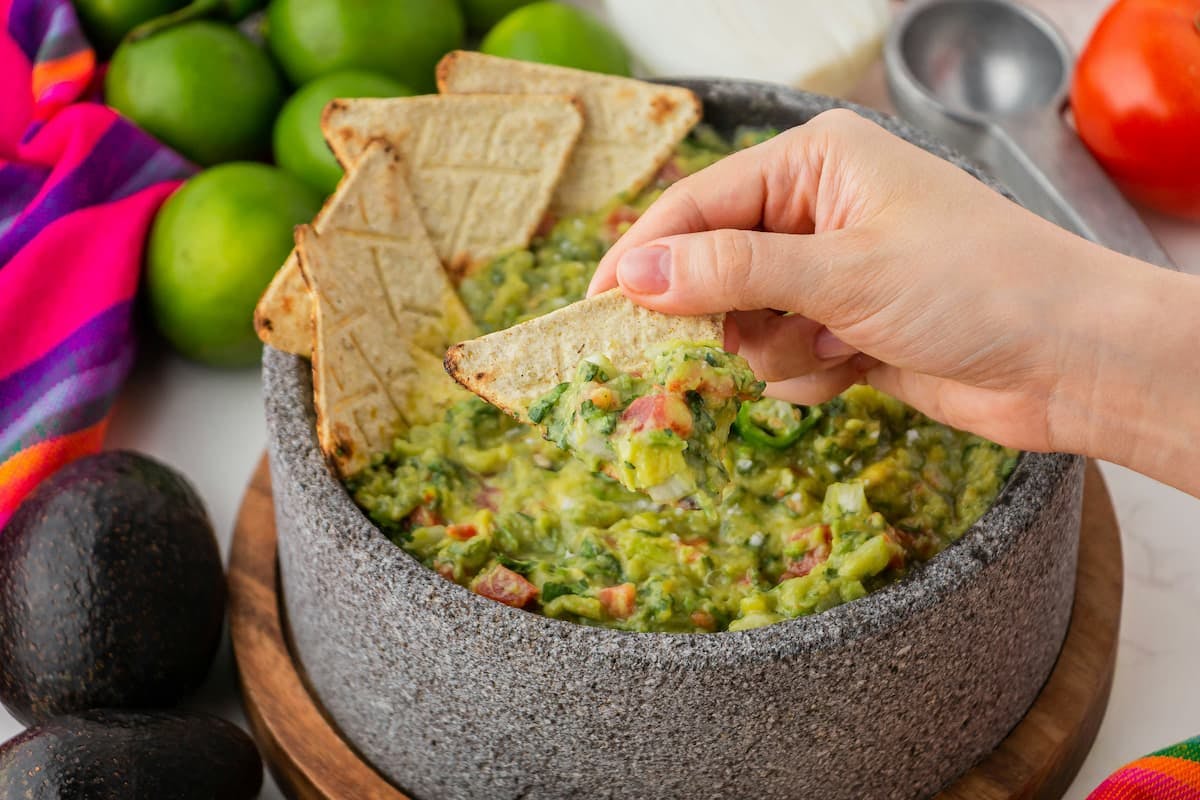Vegetarian snack featuring a tortilla chip dipped into a bowl of fresh guacamole. The stone bowl contains guacamole with chunks of tomato, onion, and cilantro, surrounded by whole avocados, limes, and a tomato on a rustic surface with a vibrant striped cloth.