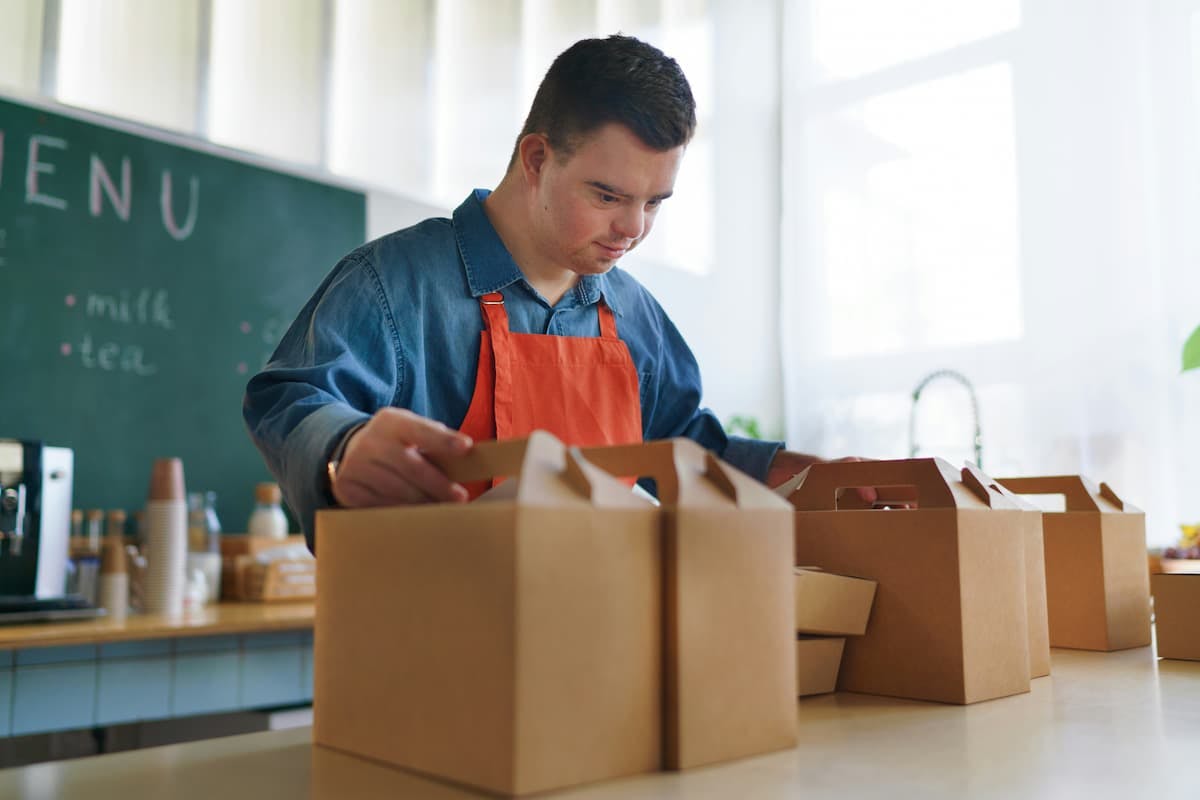 Person in a kitchen assembling food delivery boxes wearing a denim shirt and orange apron. The setup includes a chalkboard menu listing items like milk and tea, along with cups and bottles in the background, showcasing the behind-the-scenes effort of a food delivery service.