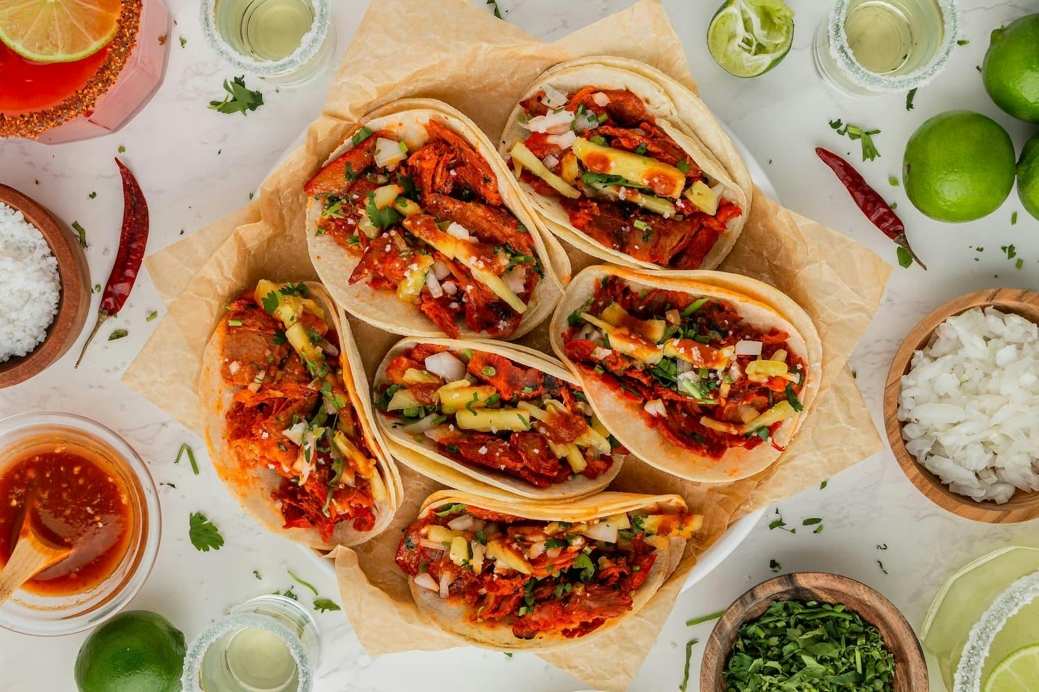 Cinco de Mayo celebration showcasing a vibrant spread of six tacos filled with juicy red meat, fresh pineapple, onions, and cilantro, set on parchment paper.