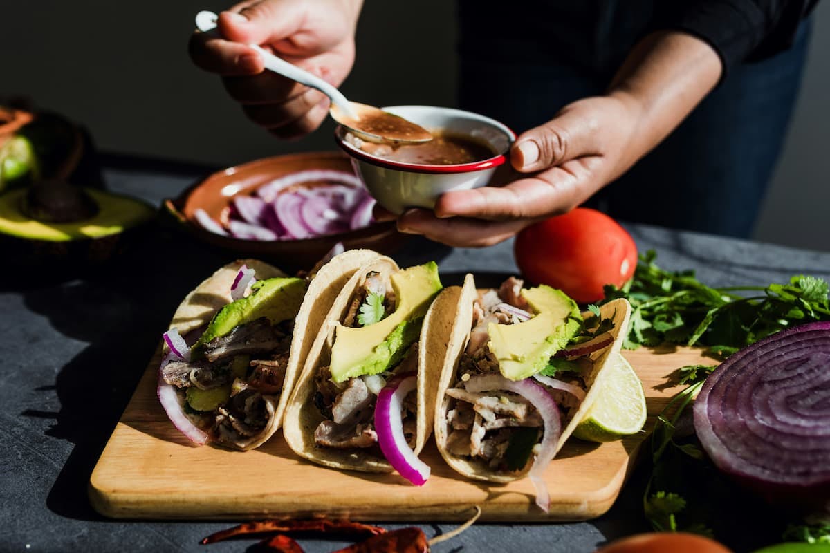 Mexican cooking class featuring a participant preparing tacos on a wooden board. The tacos are filled with seasoned meat, avocado slices, red onion, and fresh cilantro.