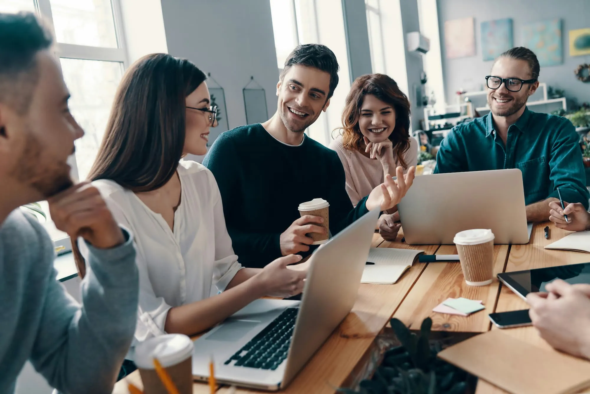 A group of people seated at a table in a modern office setting, collaborating with laptops and coffee cups. The workspace features large windows and wall decorations, emphasizing a professional and creative environment.