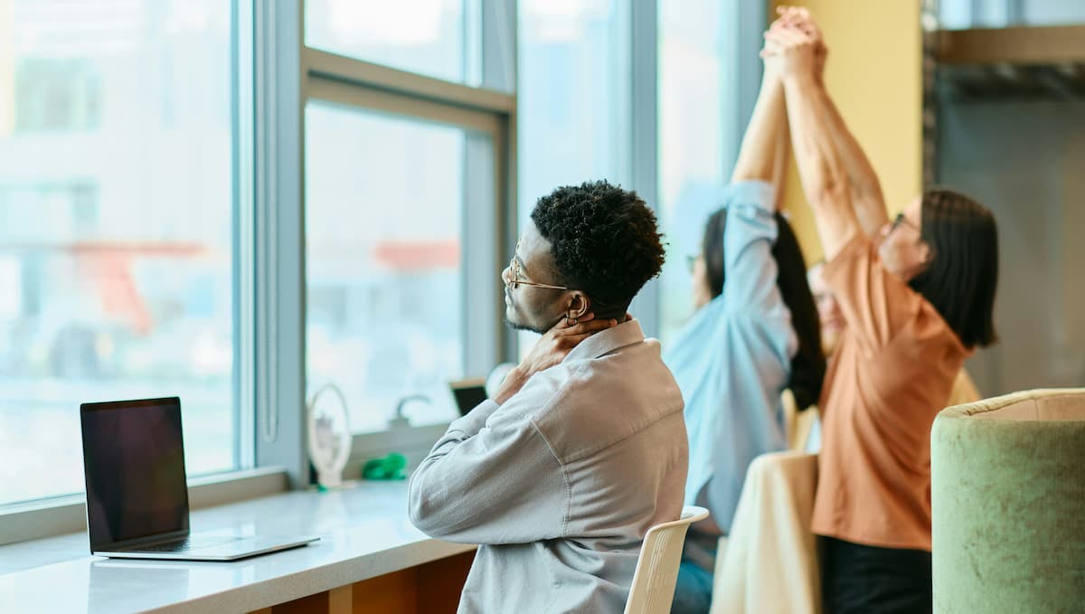 Three individuals are engaged in light stretching exercises in an office environment. One person is seated at a counter with a laptop, holding their neck gently, while two others stand and stretch their arms upward.