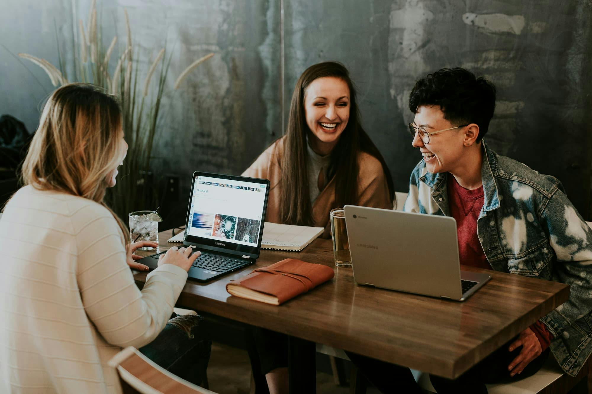 Three individuals working together at a wooden table, equipped with laptops and notebooks. The setting features a concrete wall and lush greenery in the background, creating a casual and modern workspace. Each person is engaged in their tasks, highlighting a collaborative and productive atmosphere.