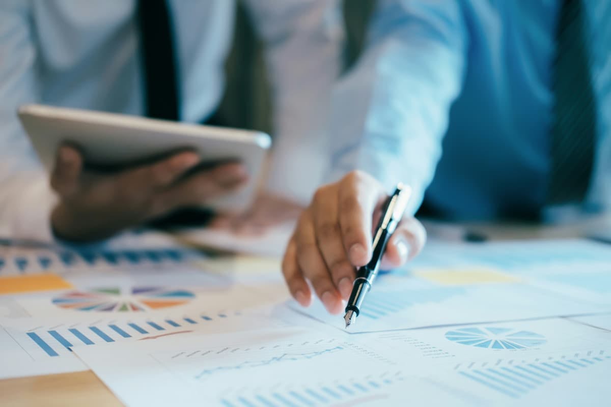 Two individuals collaborating at a desk, with one person holding a tablet and the other pointing at a document with a pen. The desk is covered with various papers featuring charts and graphs.