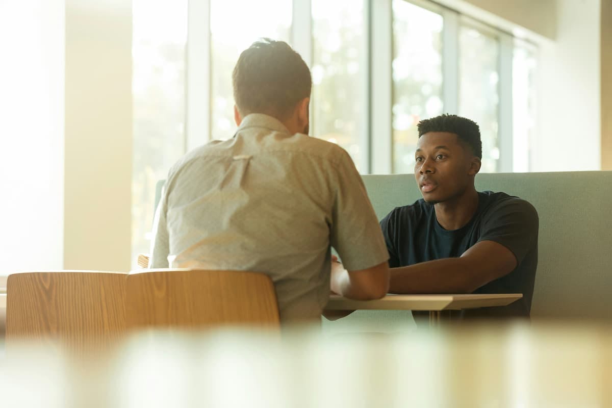 Two individuals sitting at a wooden table in a modern, well-lit room with large windows. They appear to be engaged in a conversation, with one person wearing a light-colored shirt and the other wearing a dark-colored shirt.