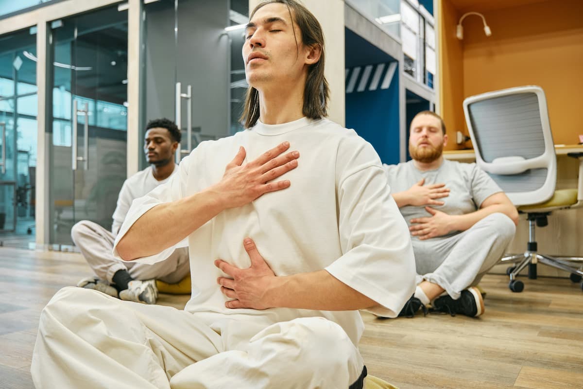 Three individuals seated on the floor of a modern office space, engaging in mindful breathing exercises. Each person places one hand on their chest and the other on their abdomen, focusing on their breath.