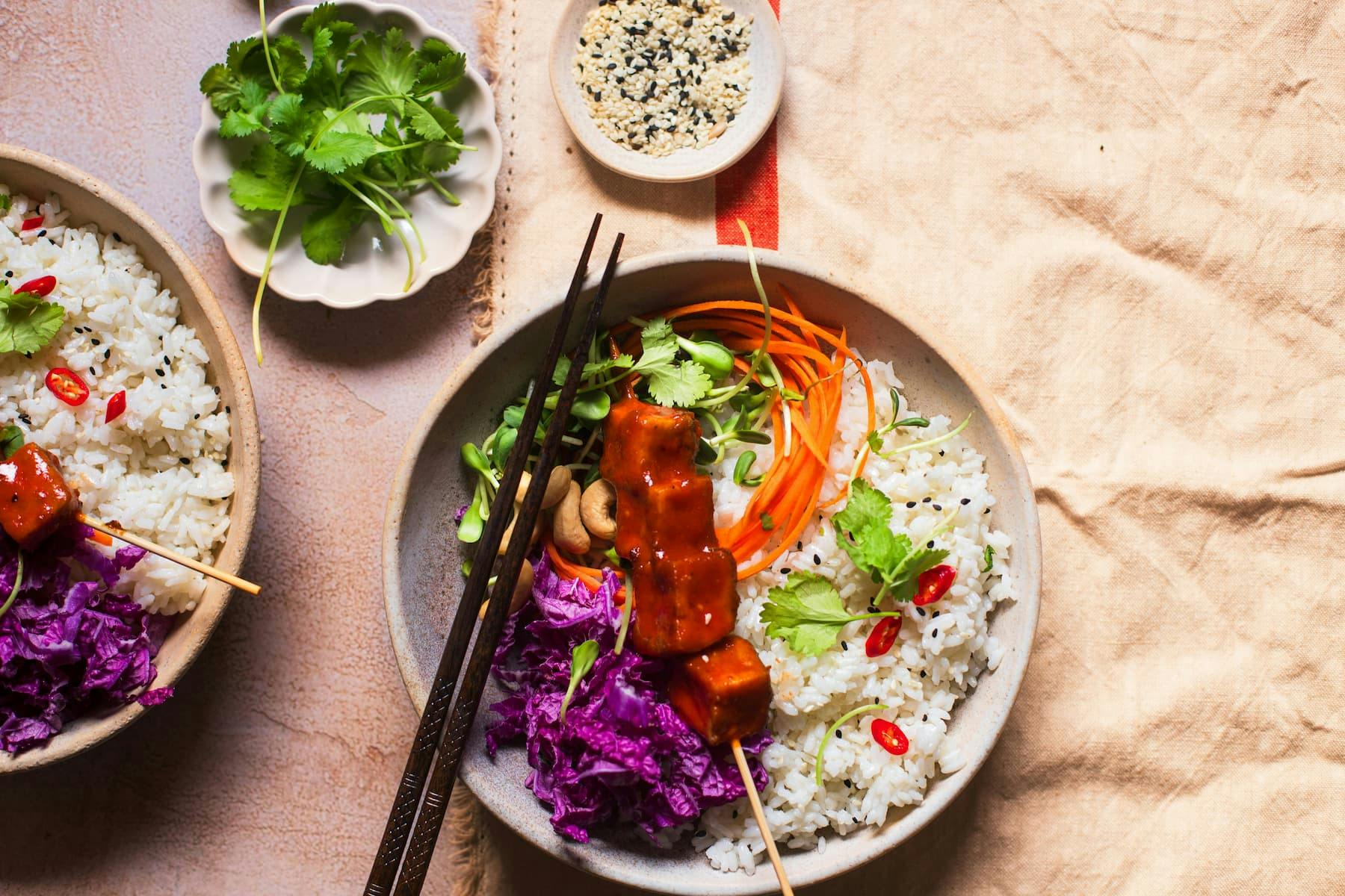 A close-up, overhead view of two bowls of vegan poke. The bowls feature fluffy white rice, vibrant purple cabbage, bright orange carrot shreds, and skewers of glazed tofu. Fresh cilantro sprigs and small slices of red chili add pops of color. A small dish of sesame seeds is also visible.