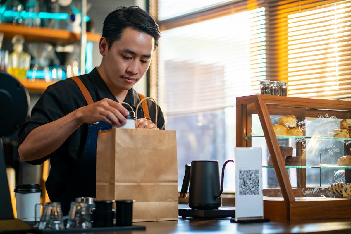In a brightly lit catering restaurant, a young man wearing a dark apron over a black shirt, gently places a small white container into a brown paper bag. He stands behind a polished counter with a black kettle and a display case showcasing baked goods. The warm sunlight streams through slatted blinds in the background.