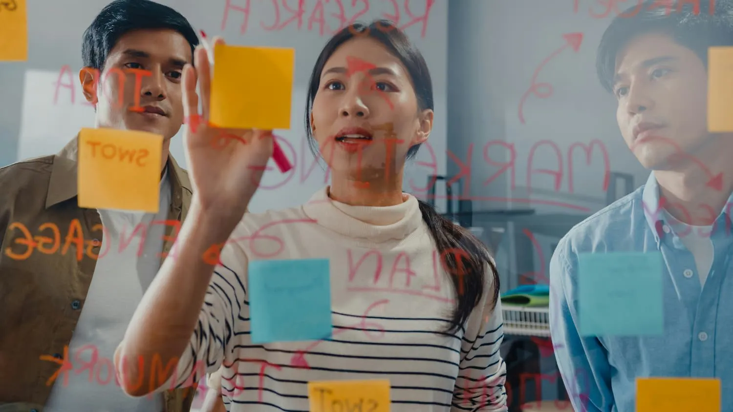 A medium shot of three people, possibly of East Asian descent, working together on a glass wall covered in sticky notes and writing, demonstrating active team participation and engagement.