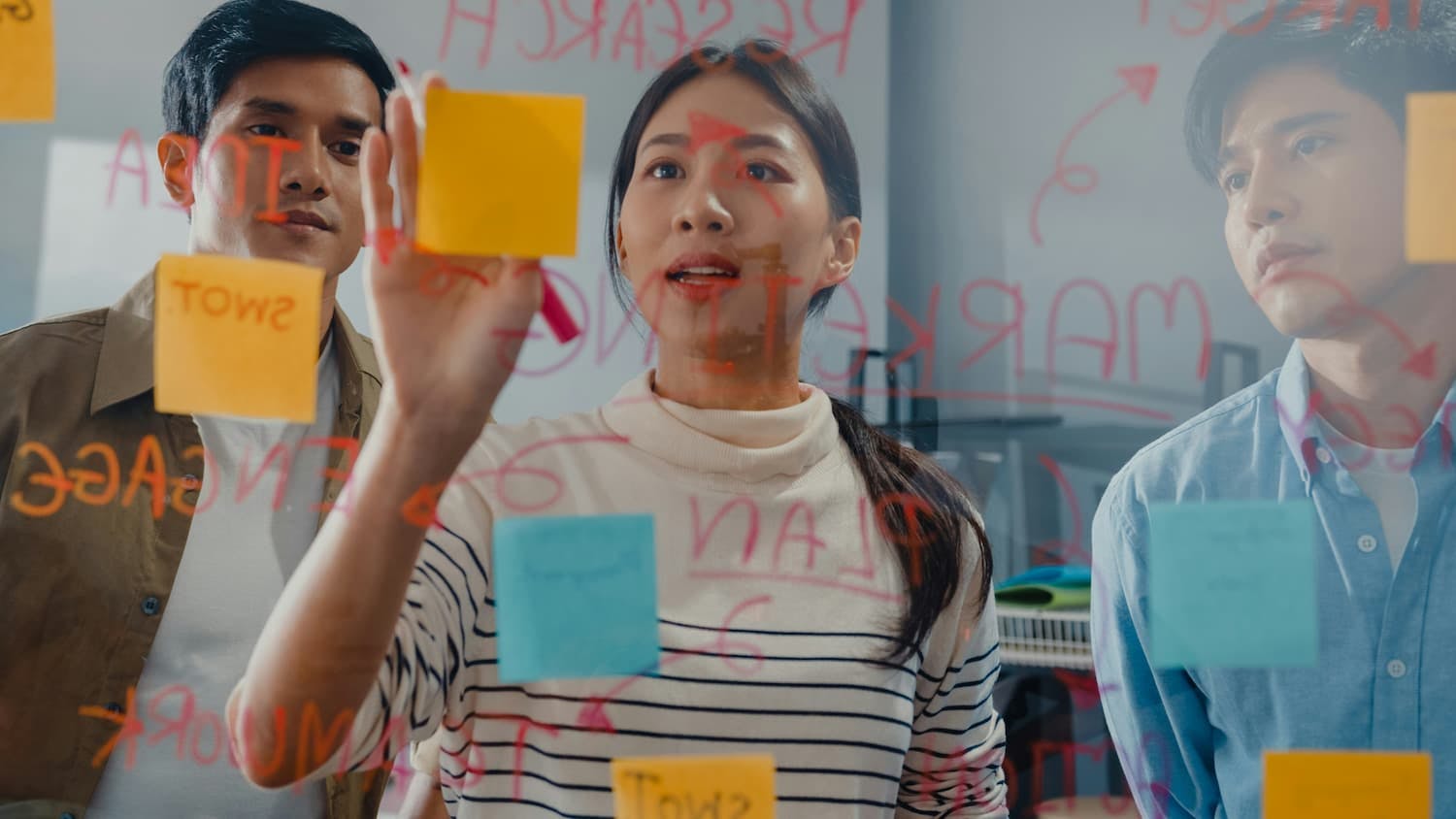 A medium shot of three people, possibly of East Asian descent, working together on a glass wall covered in sticky notes and writing, demonstrating active team participation and engagement.