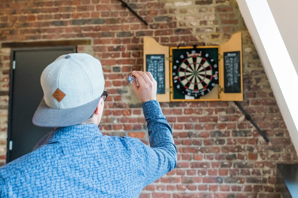 A person, possibly a male, throws a dart at a dartboard in a room with a brick wall, showcasing a recreational activity that can boost employee engagement.
