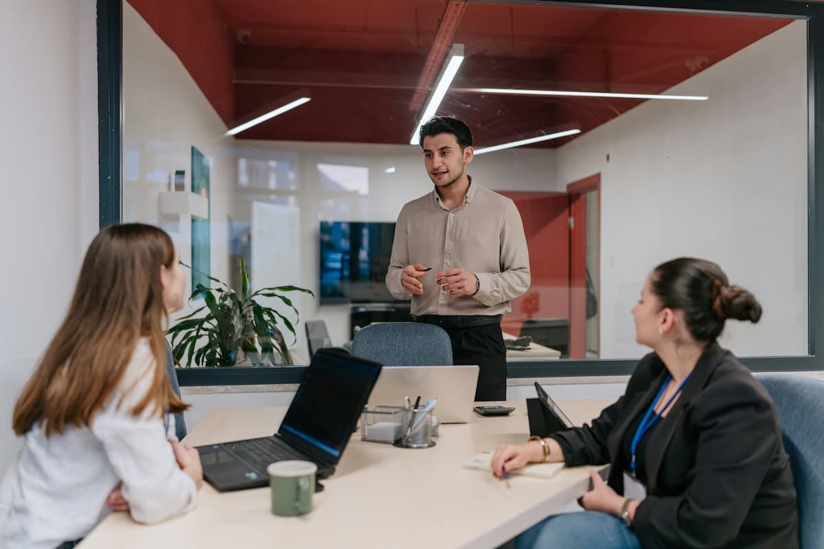 A medium shot shows three individuals, possibly two women and a man, in a modern office setting. The man stands while speaking to the two women seated at a table with laptops, suggesting an interactive meeting and emphasizing open communication as a strategy for employee engagement and retention.