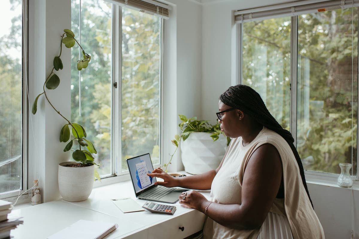 A medium shot shows a person, possibly a woman with dark hair in braids, working on a laptop at a bright desk by a large window overlooking trees.