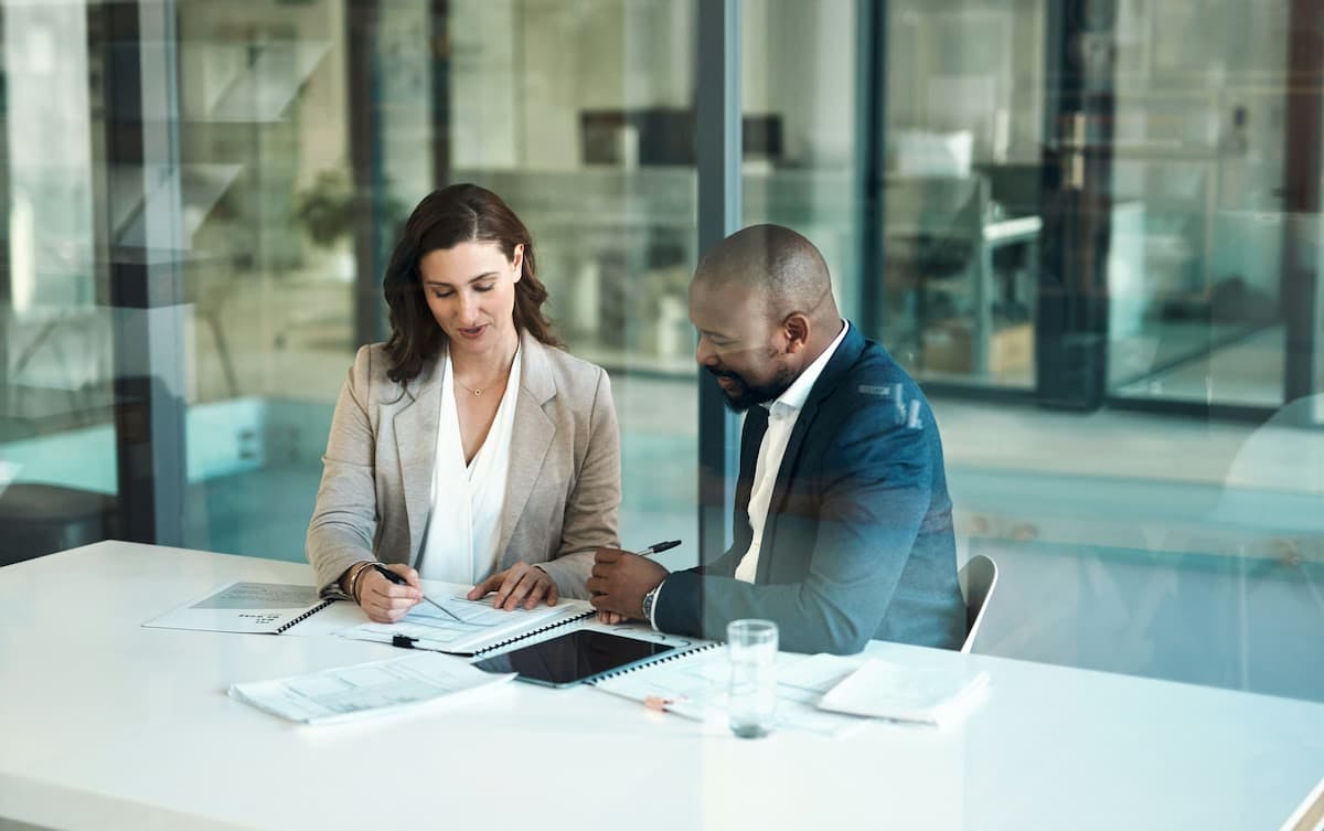 A medium shot shows a woman with light brown hair and a man with a shaved head and dark skin, working together on documents at a white table.