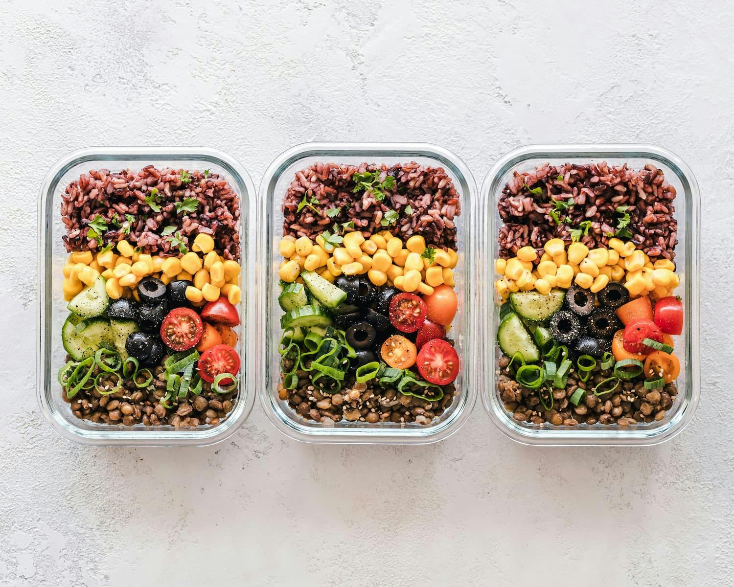 Overhead view of three glass containers with a layered box lunch: lentils, cucumbers, olives, tomatoes, corn, and red rice.