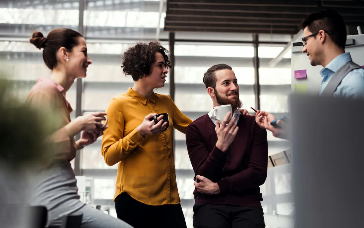 In a modern corporate dining area, a group of colleagues is gathered near a stylish coffee bar. They are enjoying coffee and engaging in conversation. The setting features large windows, sleek furniture
