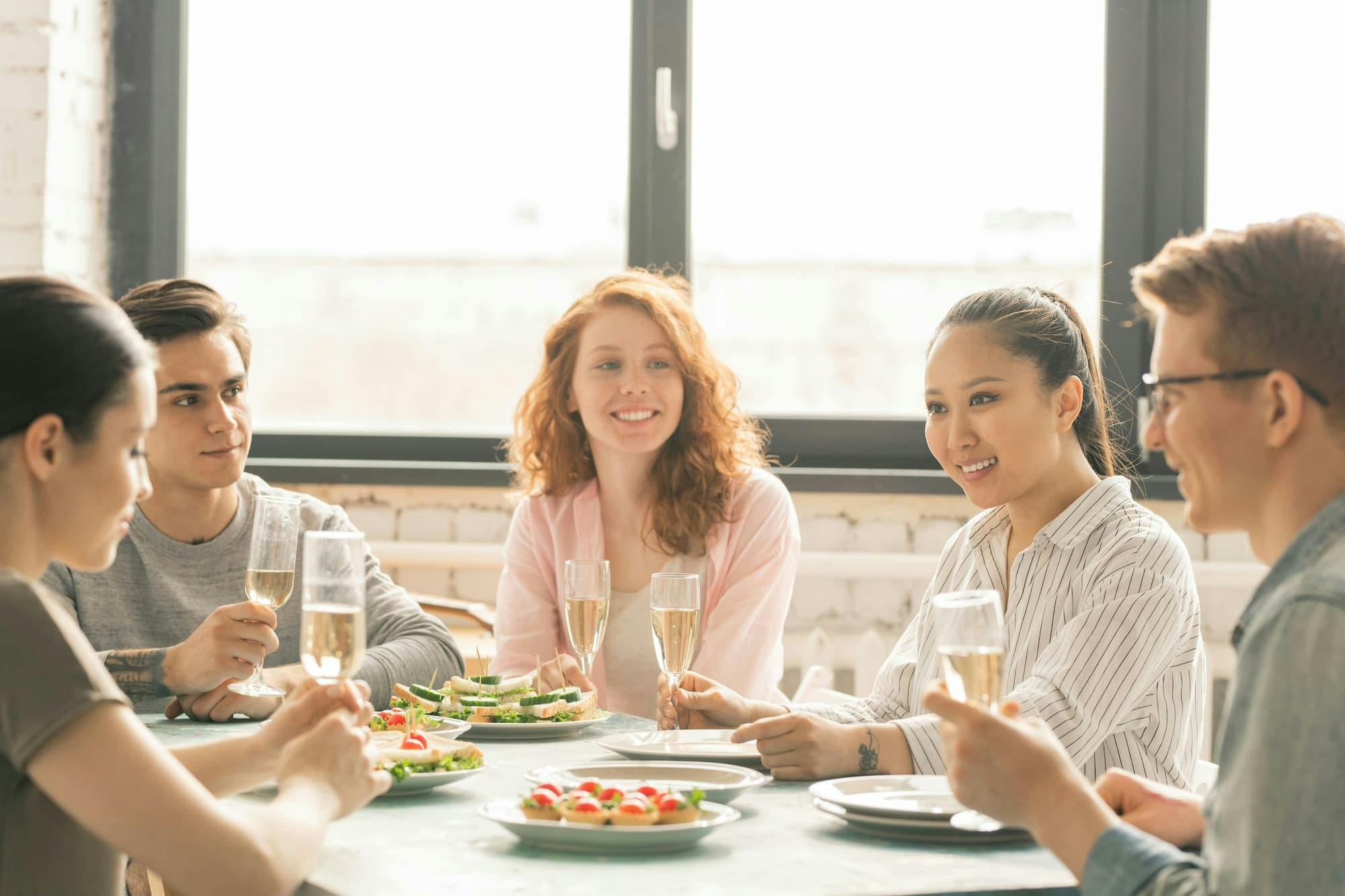 In a corporate dining space, a group of people is seated around a table, enjoying a meal together. The table is elegantly set with plates of food, including sandwiches and appetizers, while each person holds a glass of champagne.