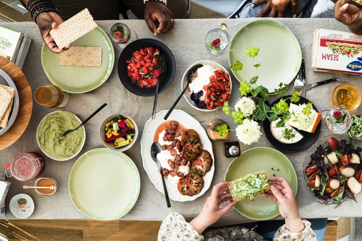 A top-down view of a dining table set for a corporate meal. The table features a variety of dishes, including sliced tomatoes, guacamole, mixed vegetables, and a bowl of strawberries. There are plates with crispbread, a jar of honey, and glasses with beverages. Several hands are visible, preparing and serving the food, evoking a sense of teamwork and collaboration in a corporate dining setting.