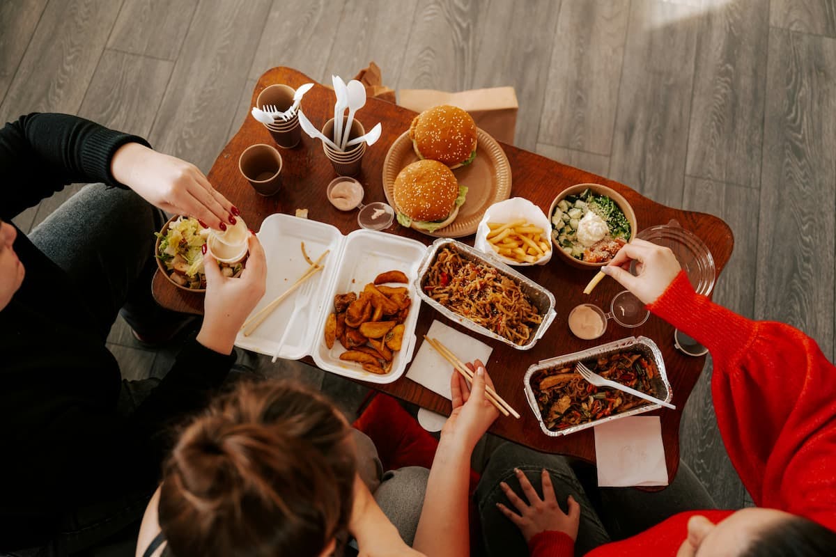 An online food delivery scene featuring a table filled with various dishes like burgers, fries, Asian noodles, and salad. Disposable utensils, cups, and sauces are scattered around, creating a casual dining setup.