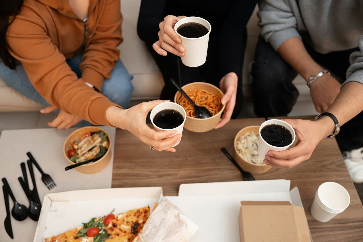 Four colleagues seated around a table in a corporate dining setting, raising cups of beverages, possibly coffee or soda, in a celebratory toast. The table is set with various dishes, including pasta, salad with grilled chicken, creamy pasta, and pizza, alongside disposable utensils and cups.