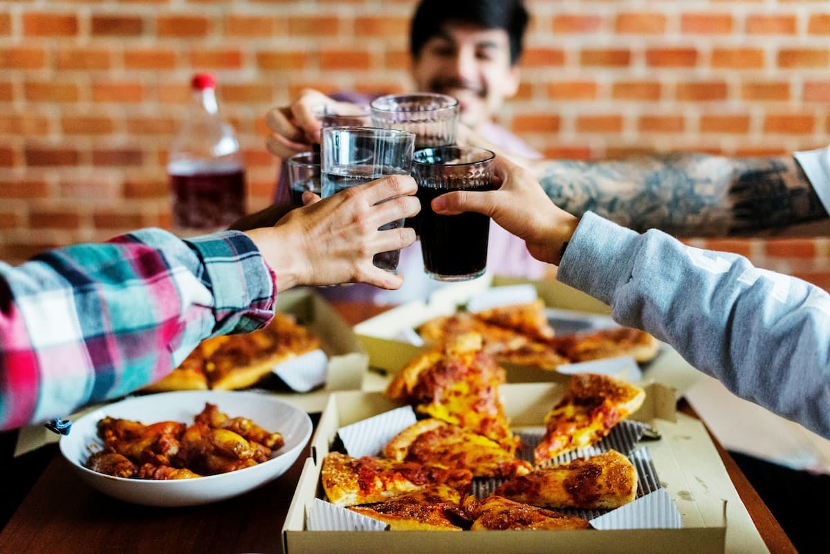 A corporate dining setup showcasing a group of colleagues gathered around a table. The table is filled with boxes of pizza and a bowl of chicken wings. The individuals are raising their glasses, filled with drinks like water and soda, in a celebratory toast. The scene captures camaraderie and shared enjoyment.