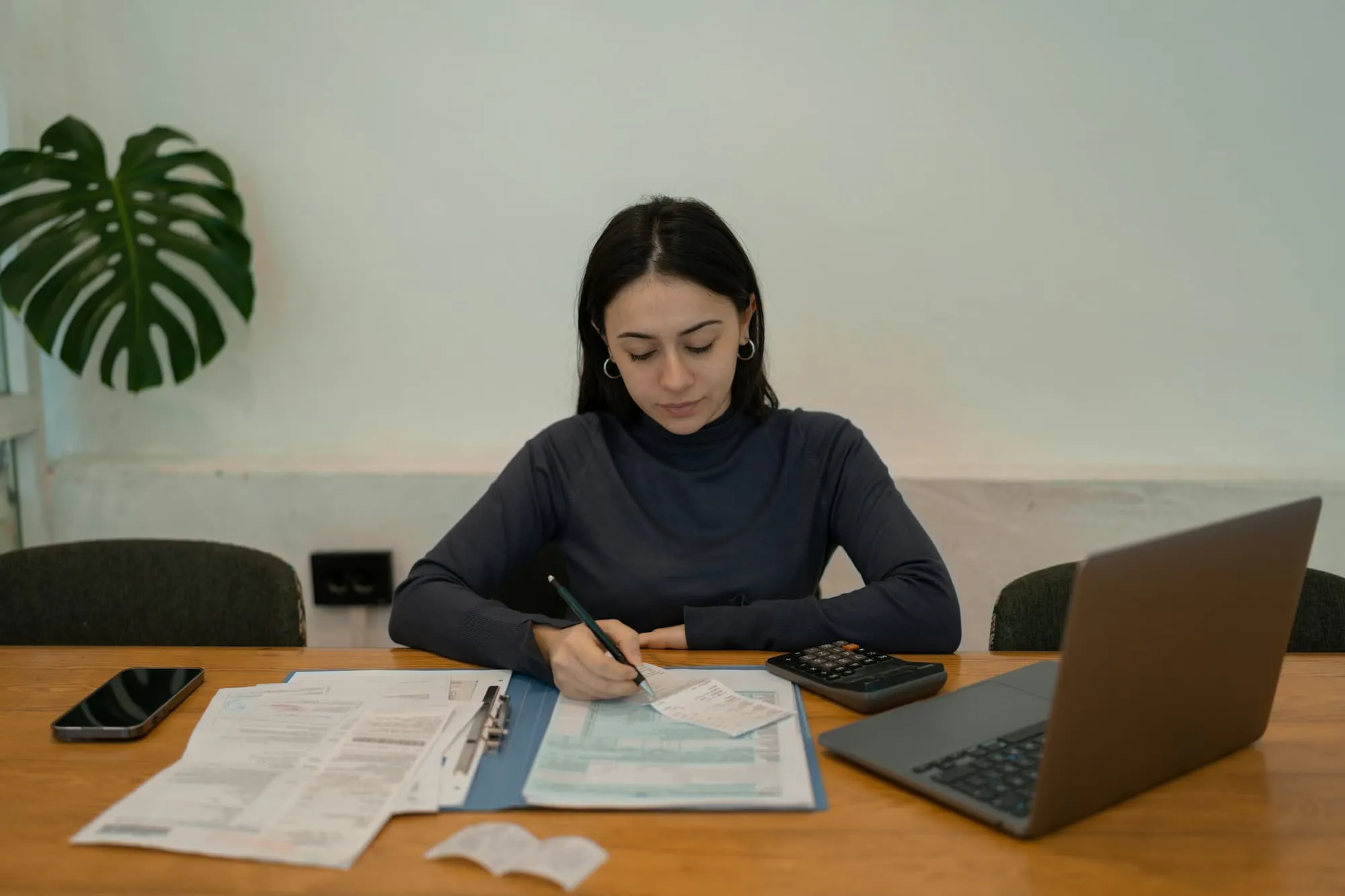 A person is seated at a wooden table, working on financial documents. The table features items such as a smartphone, a calculator, a laptop, and several sheets of paper. The person is writing on a document with a pen, surrounded by receipts and other paperwork.