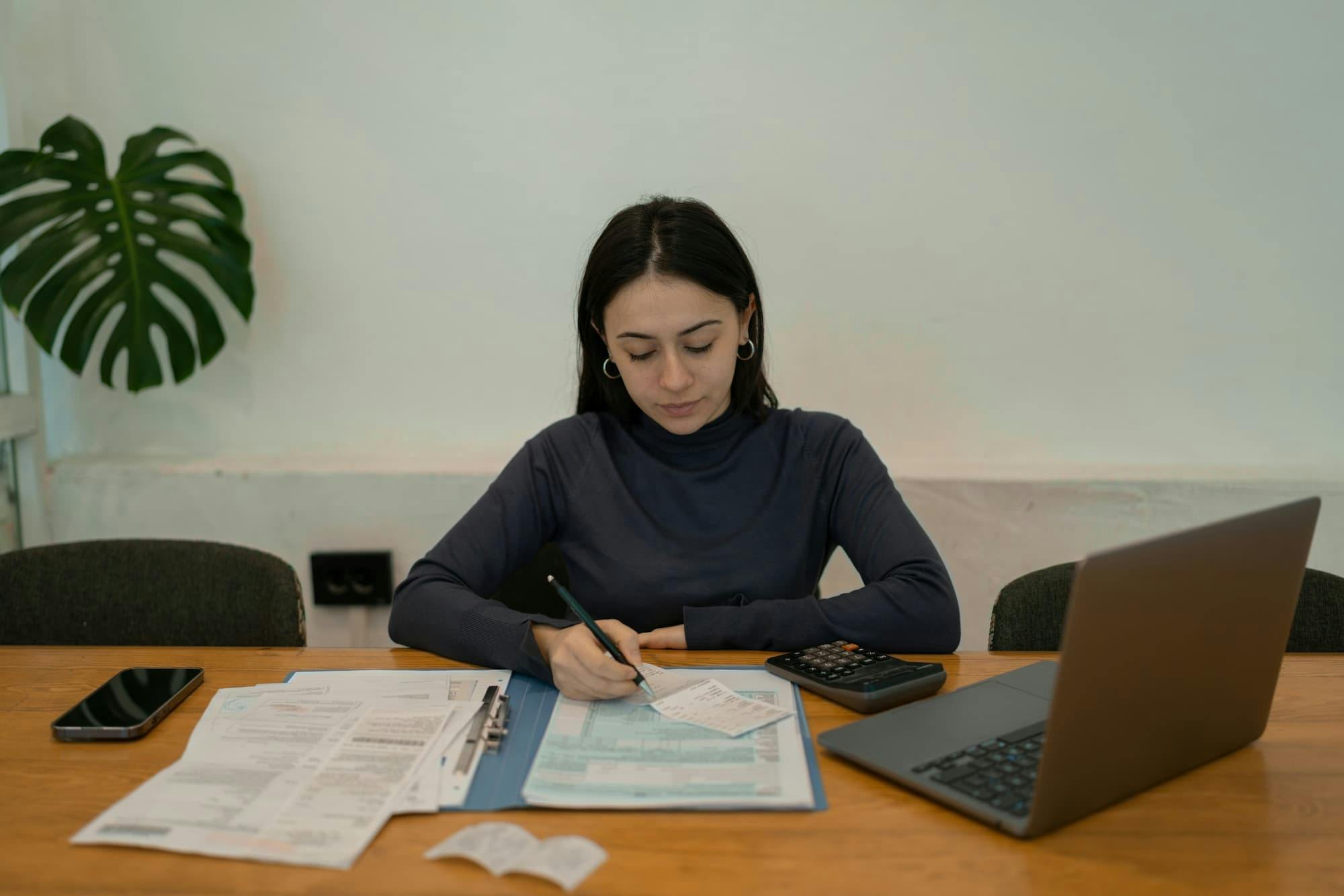 A person is seated at a wooden table, working on financial documents. The table features items such as a smartphone, a calculator, a laptop, and several sheets of paper. The person is writing on a document with a pen, surrounded by receipts and other paperwork.