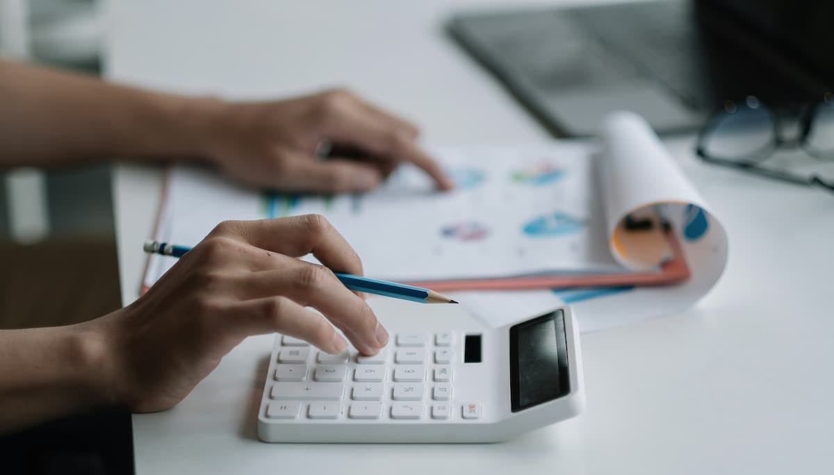 Person using a white calculator while holding a pencil, with a document featuring colorful charts and graphs on the desk. The image represents the process of analyzing and calculating costs associated with employee turnover