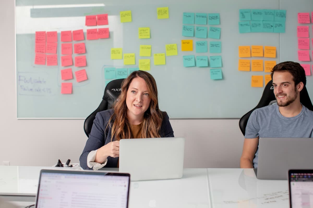 Two employees sit at a conference table with laptops, engaged in discussion. Behind them, a whiteboard covered with colorful sticky notes suggests brainstorming or planning sessions. This image highlights a collaborative workplace environment, which can be impacted by the costs and challenges of frequent employee turnover.