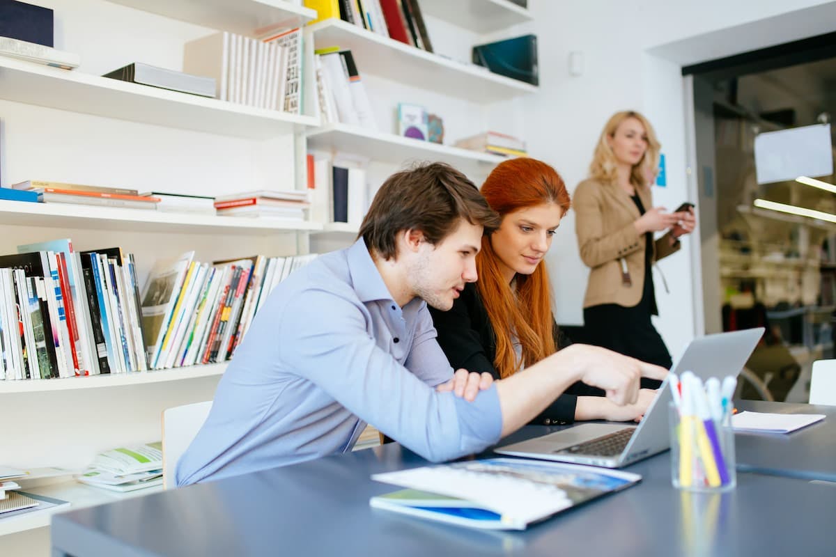 Group of people collaborating in a modern office. Two individuals are working on a laptop at a table, surrounded by bookshelves filled with documents. Another person in the background is using a smartphone.
