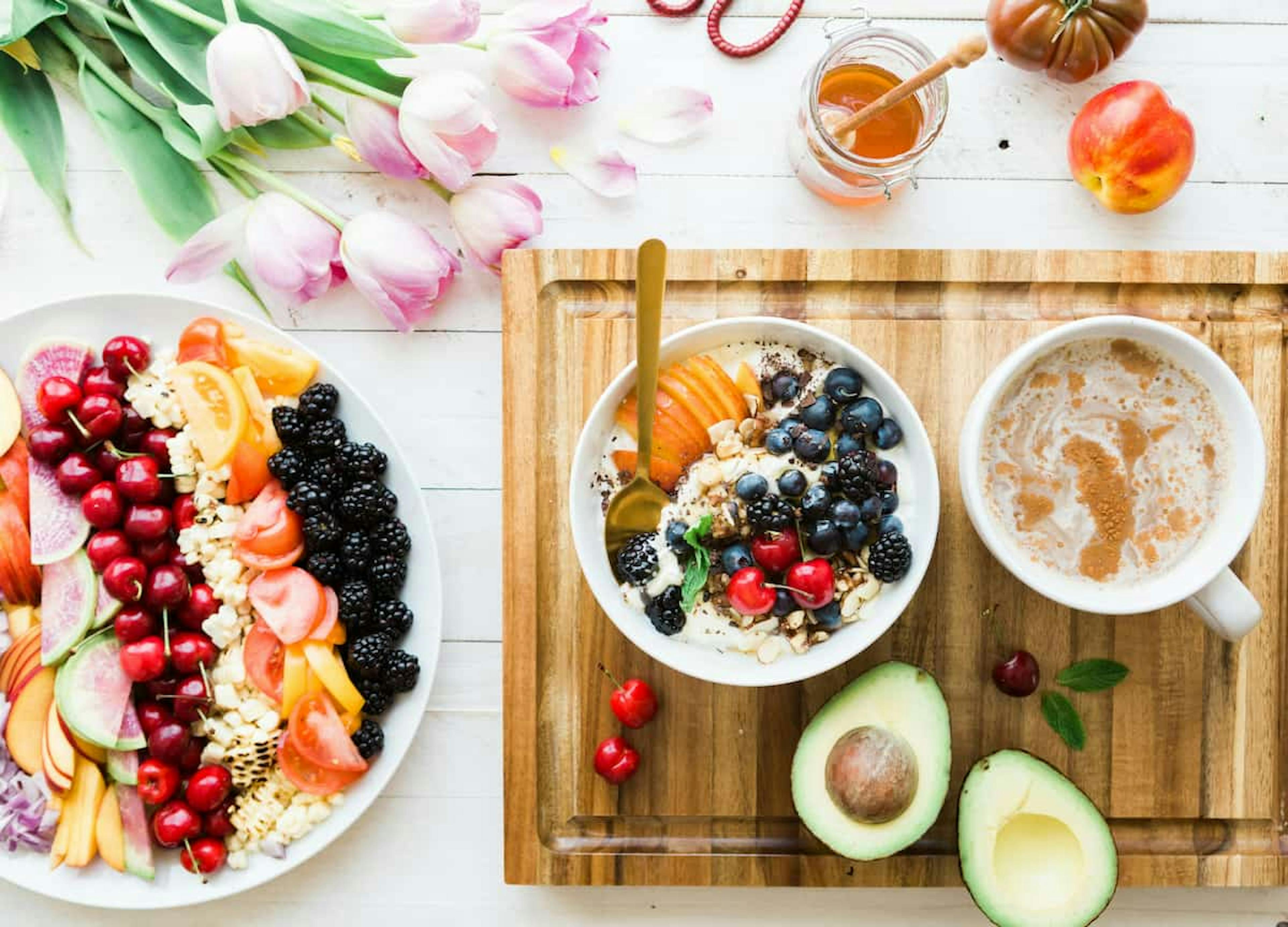 A vibrant breakfast setup featuring a plate of fresh fruits and vegetables, a bowl of yogurt topped with berries and nuts, a cup of coffee, and a halved avocado, arranged on a wooden tray alongside tulips and a jar of honey.