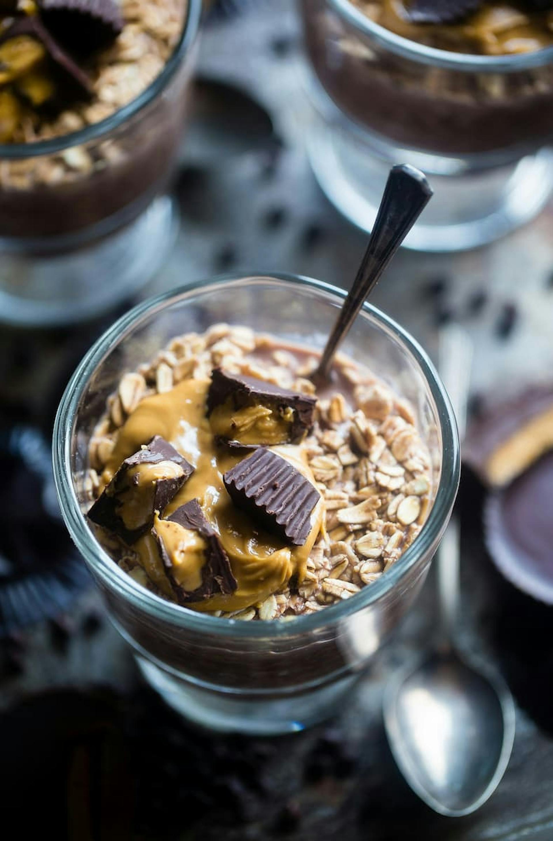 A glass filled with overnight oats layered with chocolate, creamy peanut butter, and topped with chunks of peanut butter cups, served with a spoon on a dark textured surface.