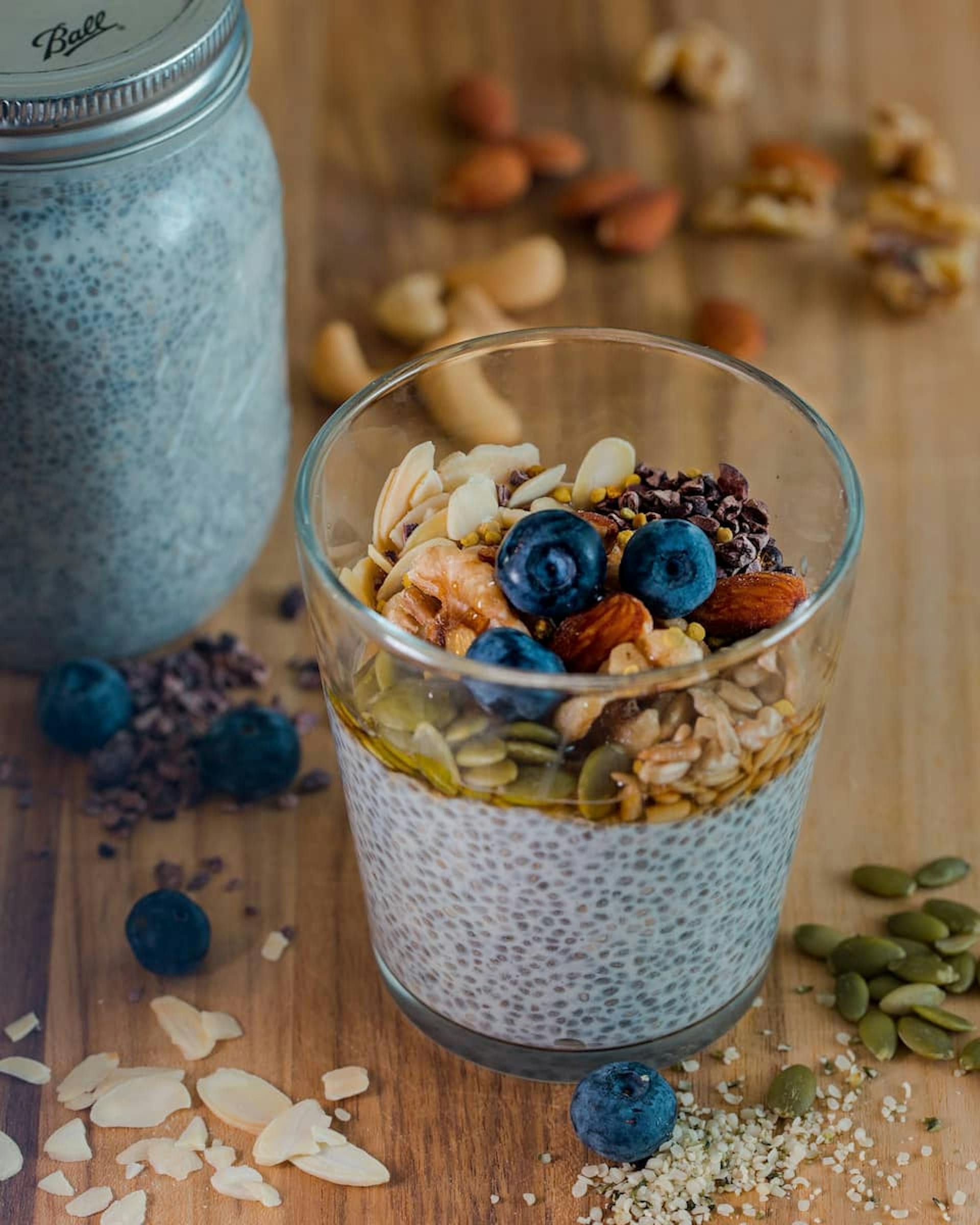 A glass of chia pudding topped with fresh blueberries, nuts, seeds, and a drizzle of honey, placed on a wooden surface with scattered almonds, walnuts, and cacao nibs in the background. A jar of chia pudding is also visible.