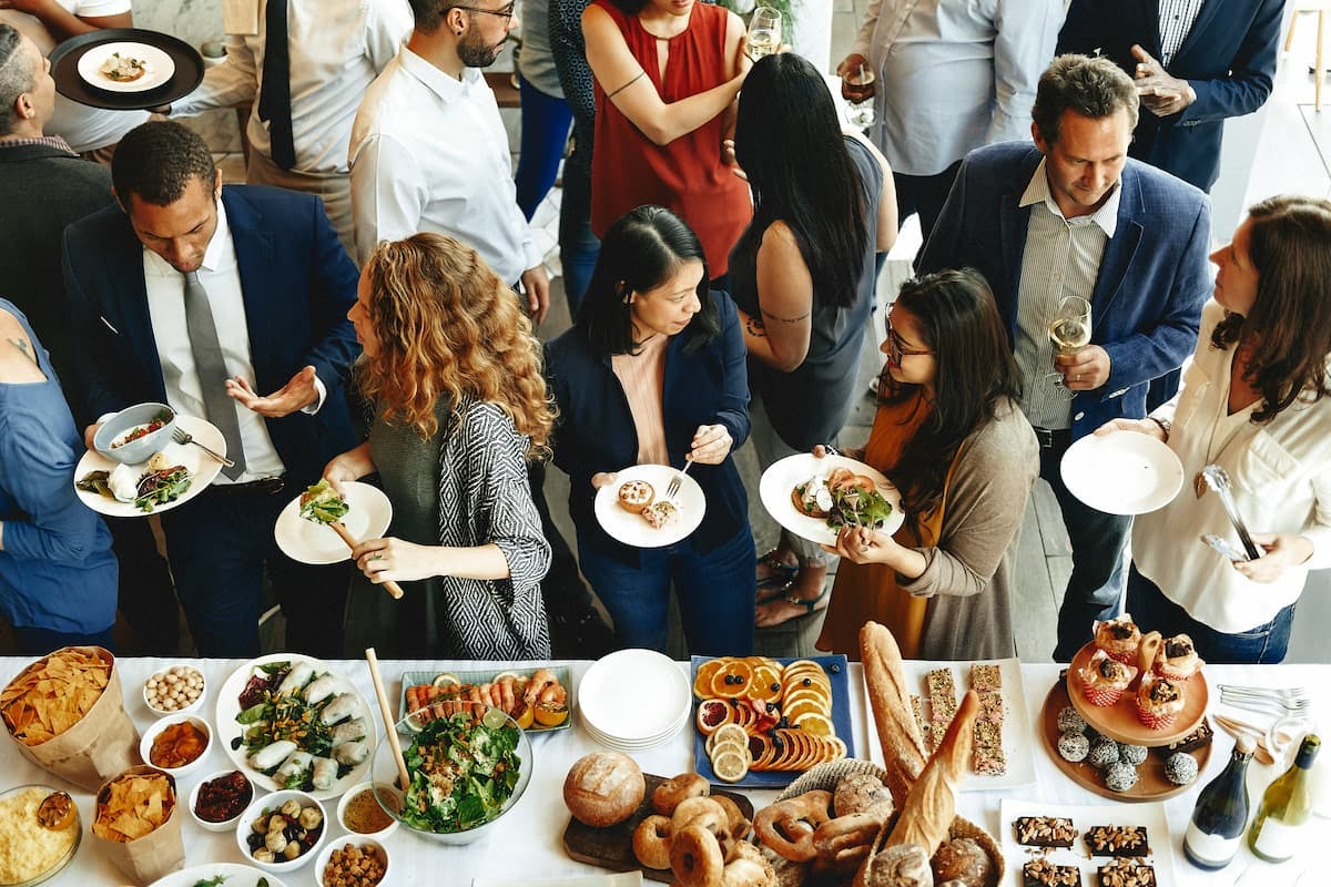 People at a social event chatting and helping themselves from a table full of a variety of foods, including salads, breads, and gourmet dishes