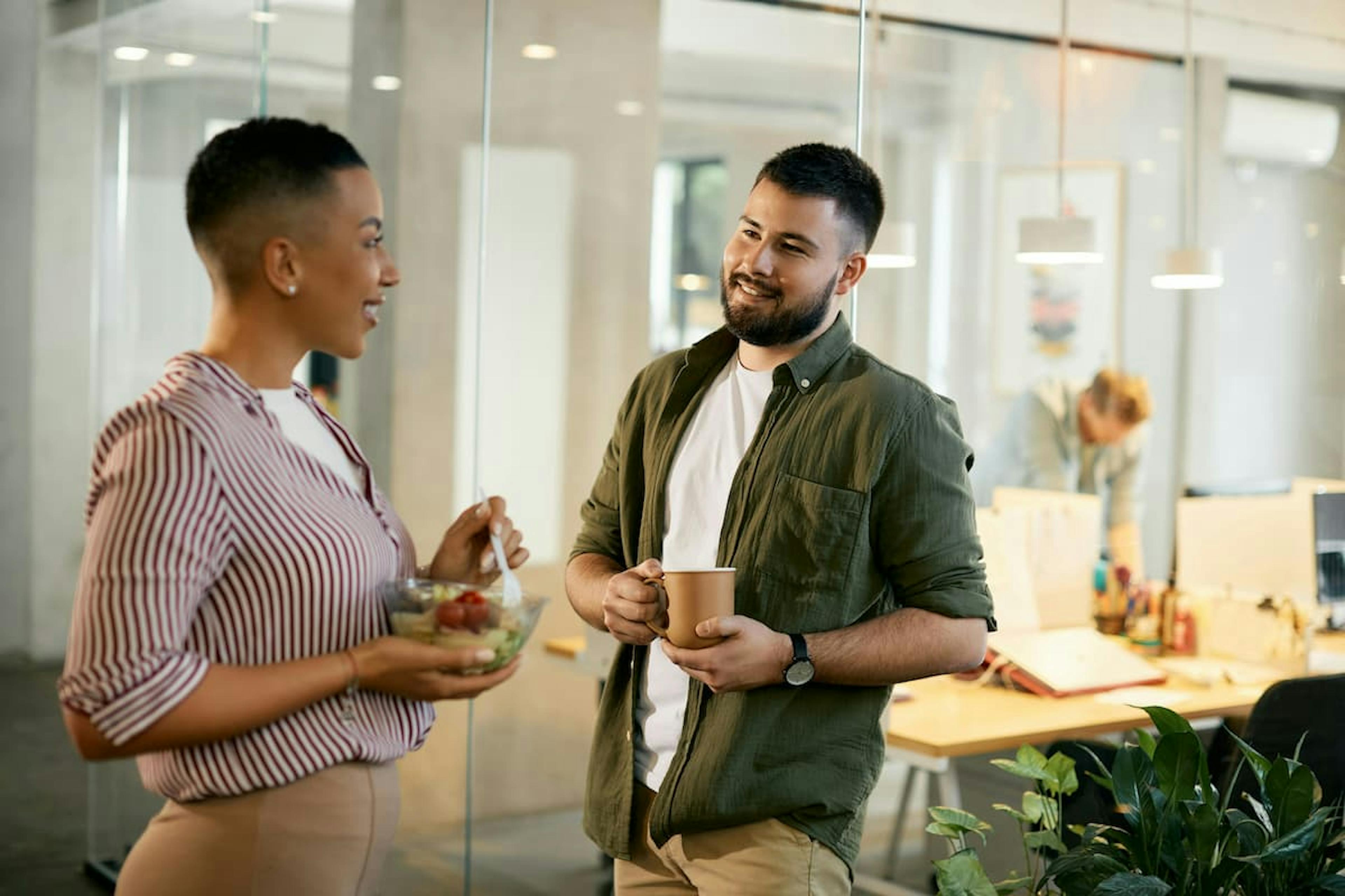 A woman and a man talking in a modern office. The woman is holding a bowl of salad, and the man has a cup of coffee