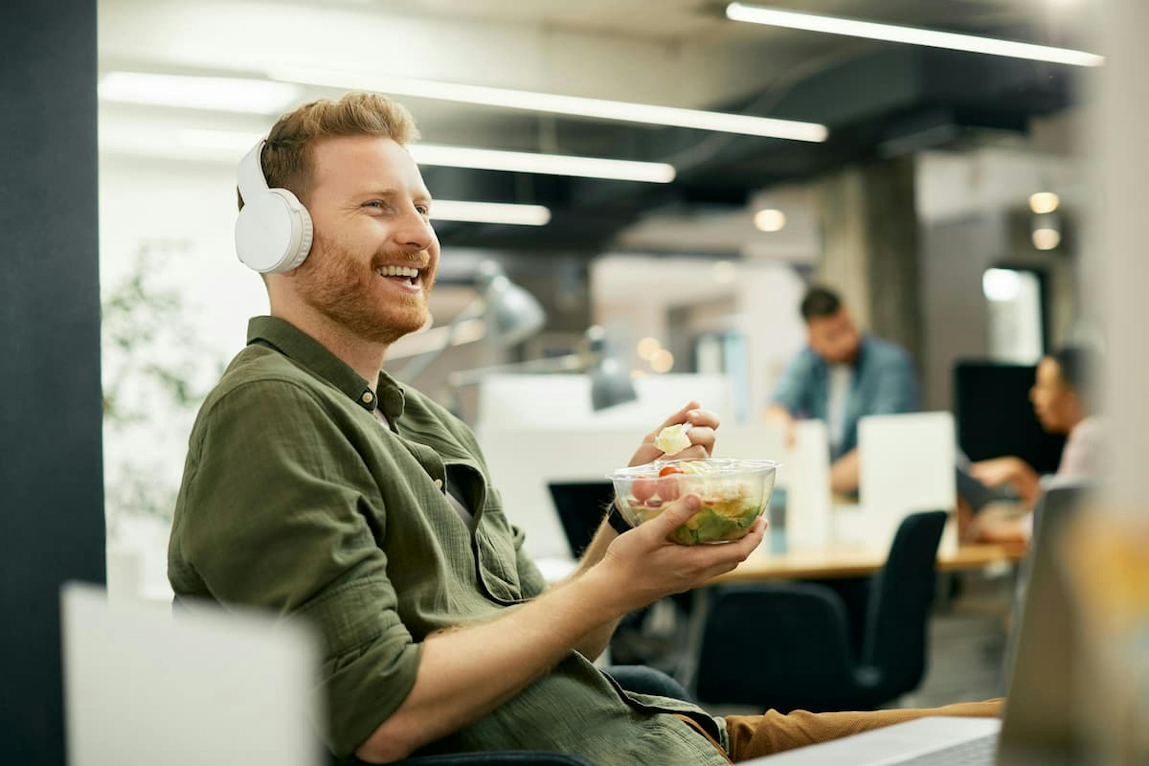 A man wearing headphones smiling while eating a salad in a modern office