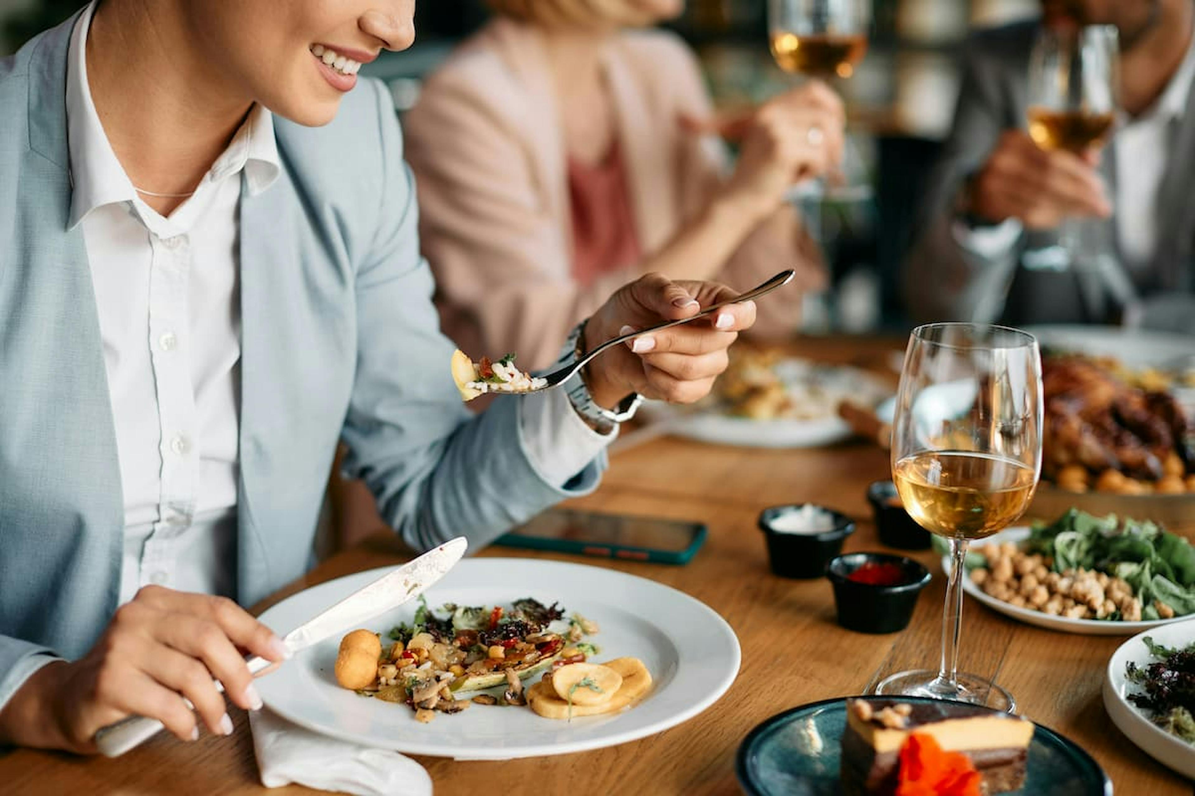 A woman smiling while eating at a dinner with friends, with dishes of food and wine glasses on the table