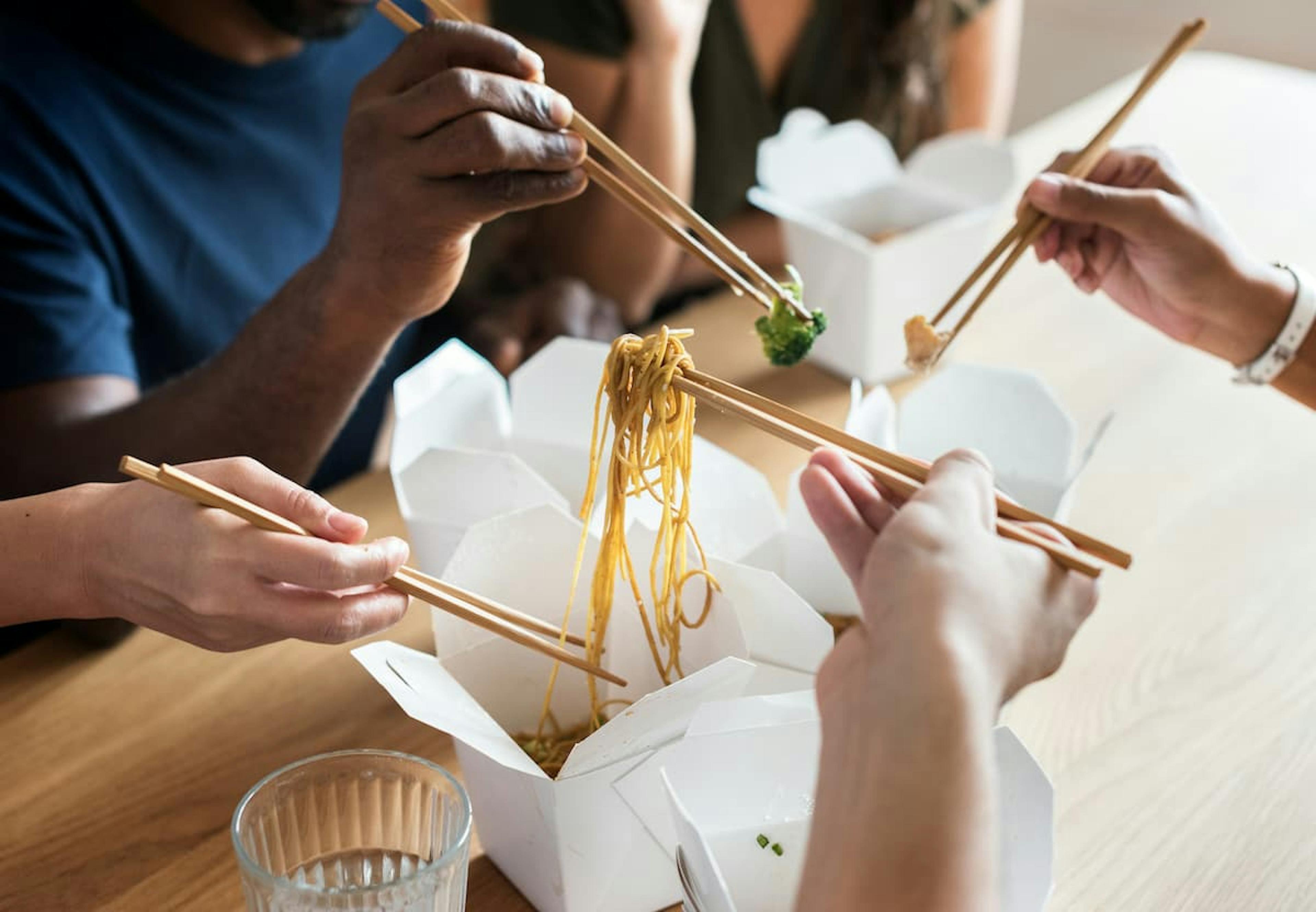 A group of people sharing food in white boxes, using chopsticks to pick up noodles and vegetables