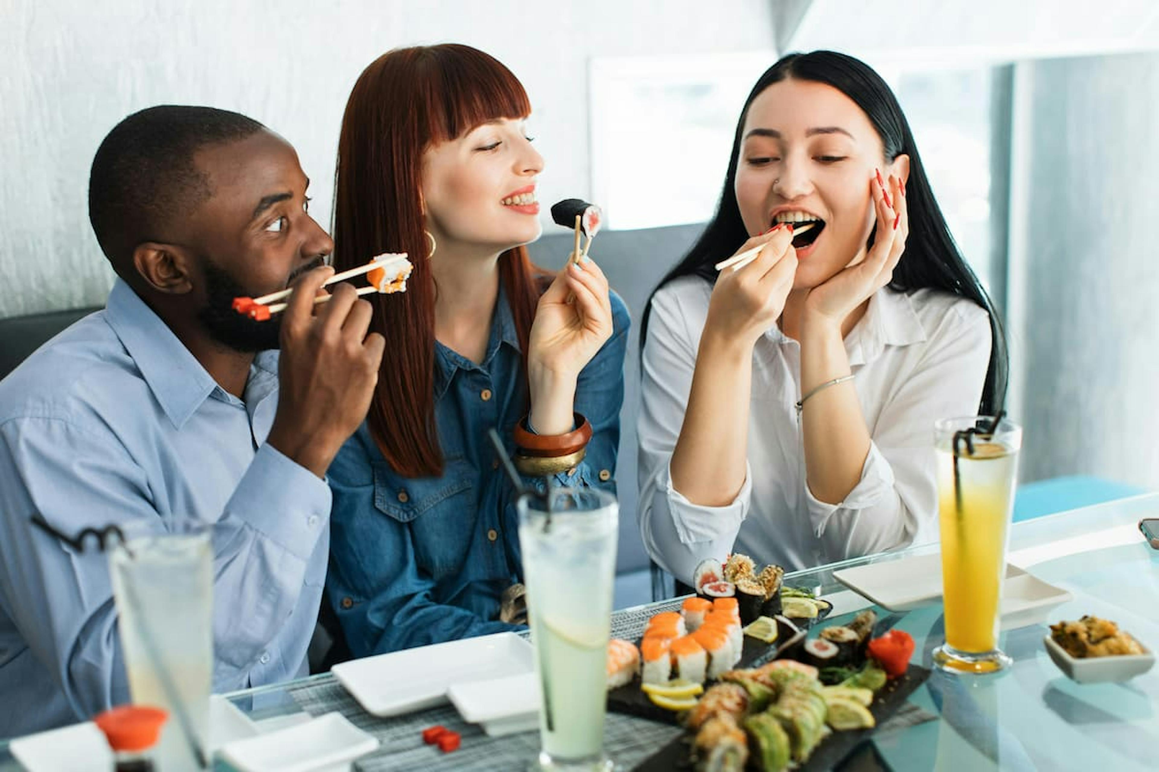 Three friends having fun in the office, enjoying sushi with chopsticks, accompanied by drinks and various sushi dishes