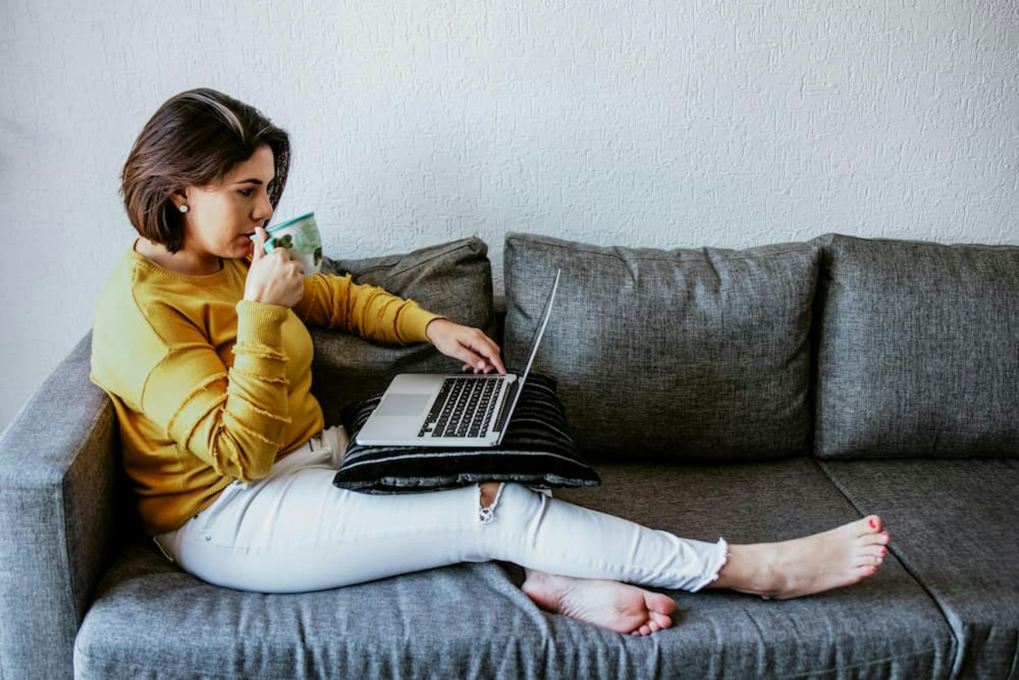 Woman sitting on a sofa, using a laptop and holding a drink, wearing a yellow sweater and white pants. Cozy and modern environment