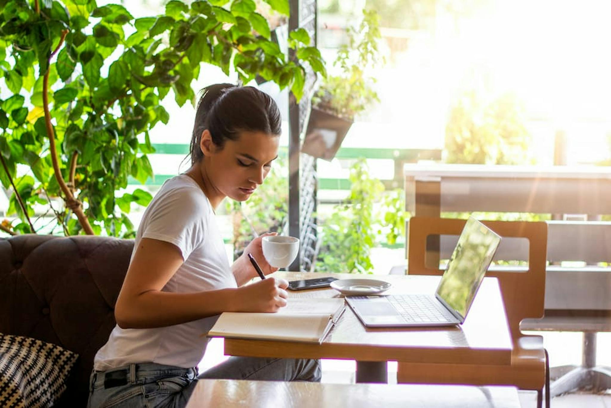 Woman working at the office environment, writing in a notebook while enjoying a cup of coffee. A laptop and a smartphone are on the table