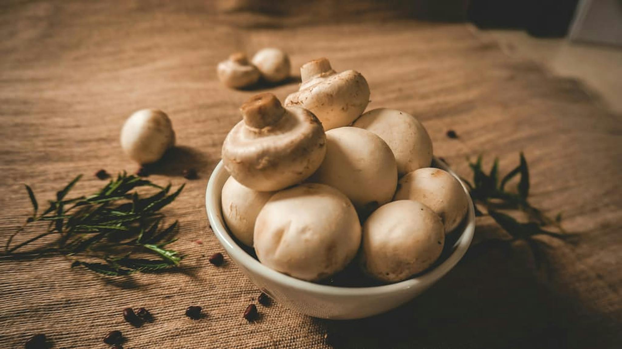 A bowl full of fresh mushrooms on a rustic table, with some herbs around