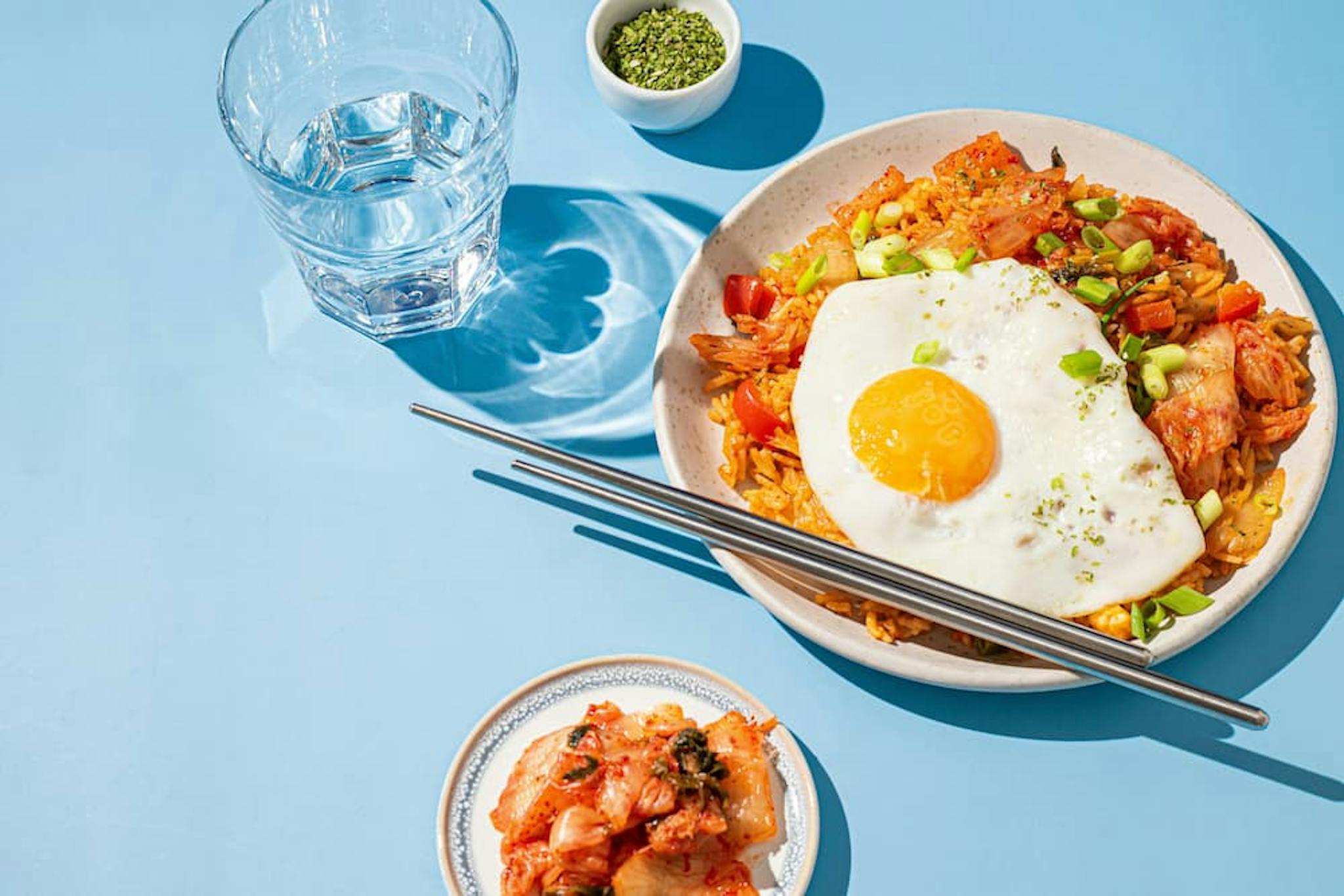 Colorful dish with fried rice, sunny-side up egg, and kimchi, accompanied by a glass of water and green powder.