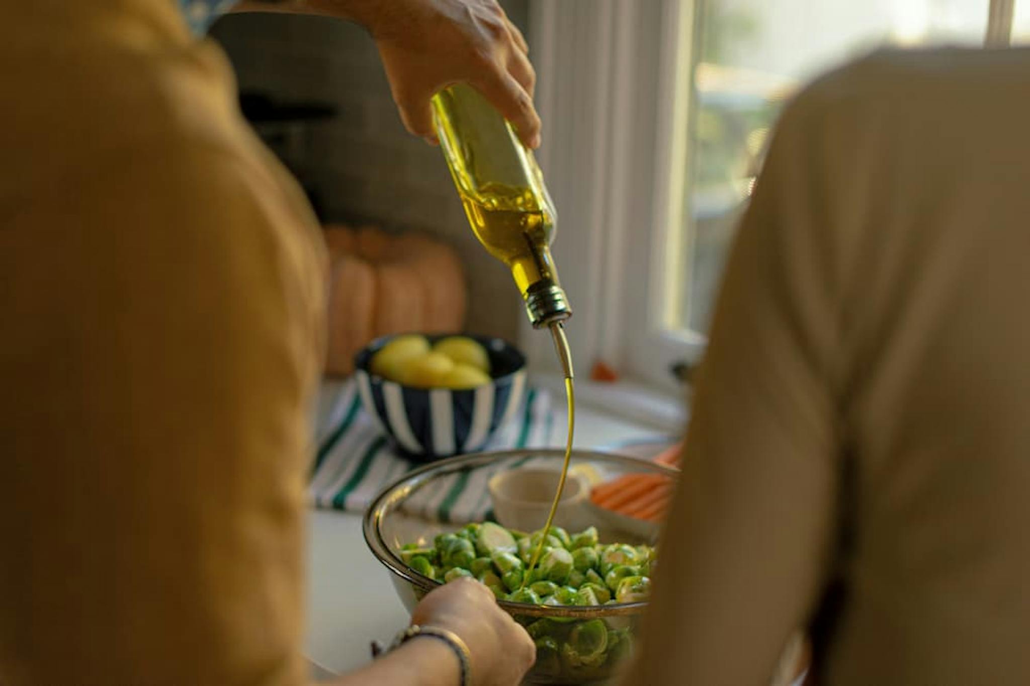 Two people in the kitchen preparing a healthy dish, with one of them pouring olive oil into a bowl with vegetables