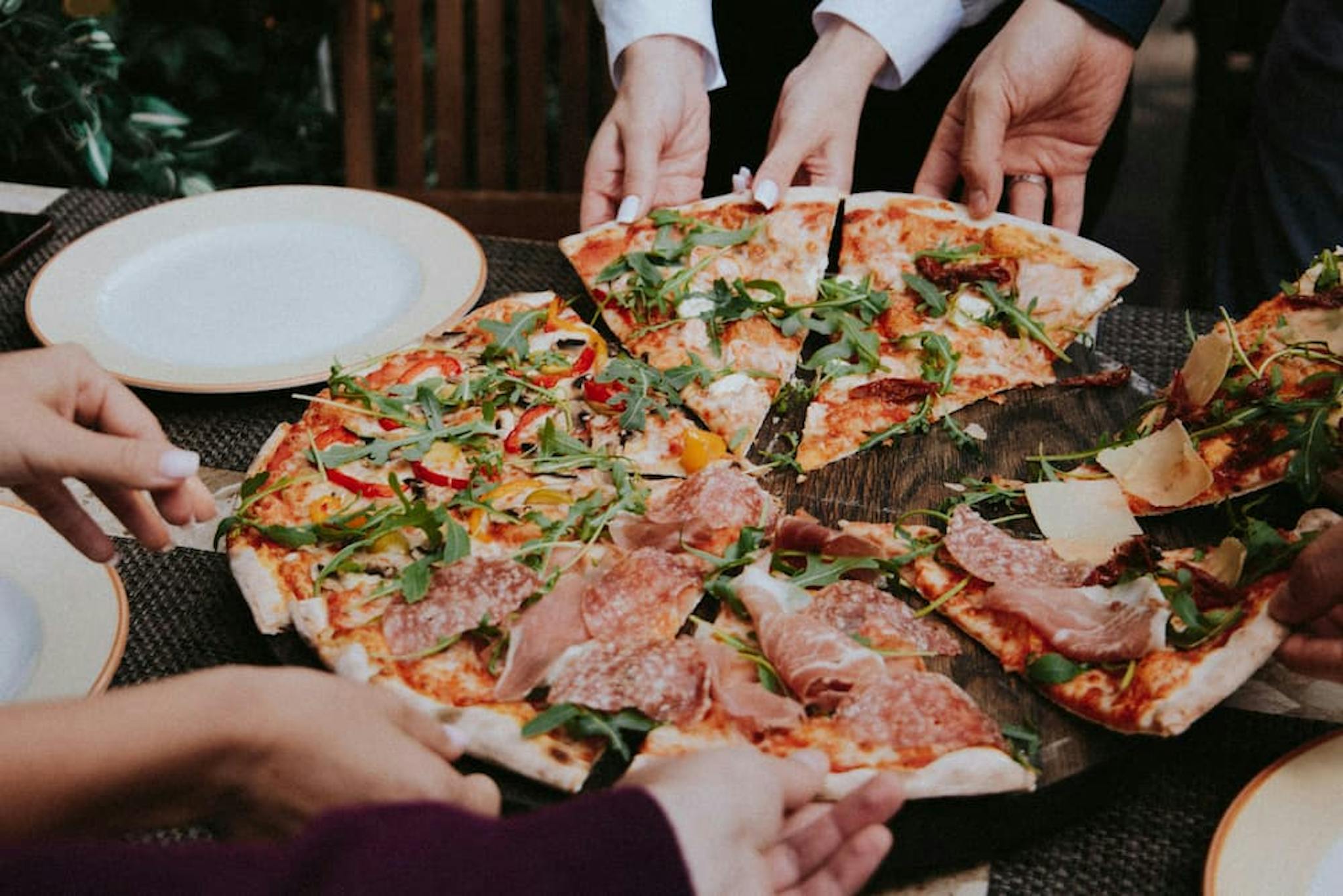 Slices of pizza being served on a table, with various fresh ingredients and a group of people sharing the meal