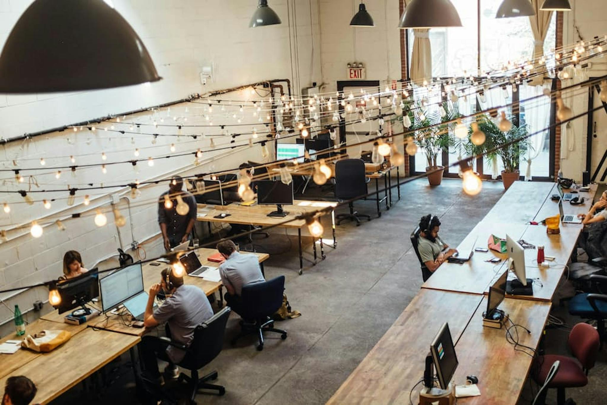 Modern office environment with pendant lights, work tables, and people using computers in a collaborative space