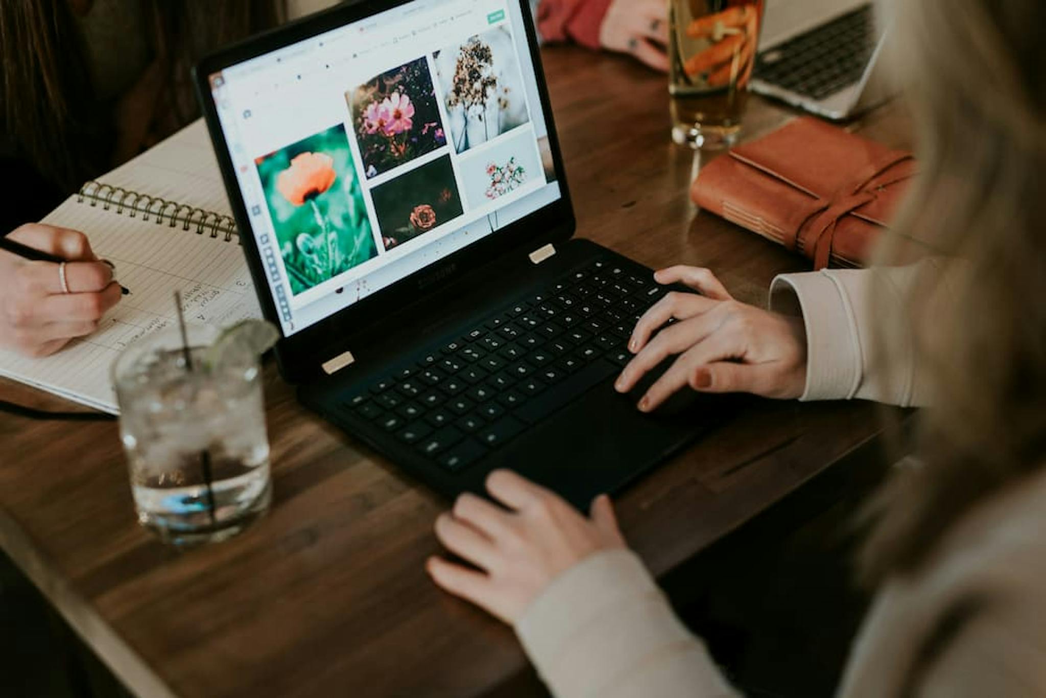 A person using a laptop on a wooden table, searching for images of flowers, with notes and a glass of water beside them