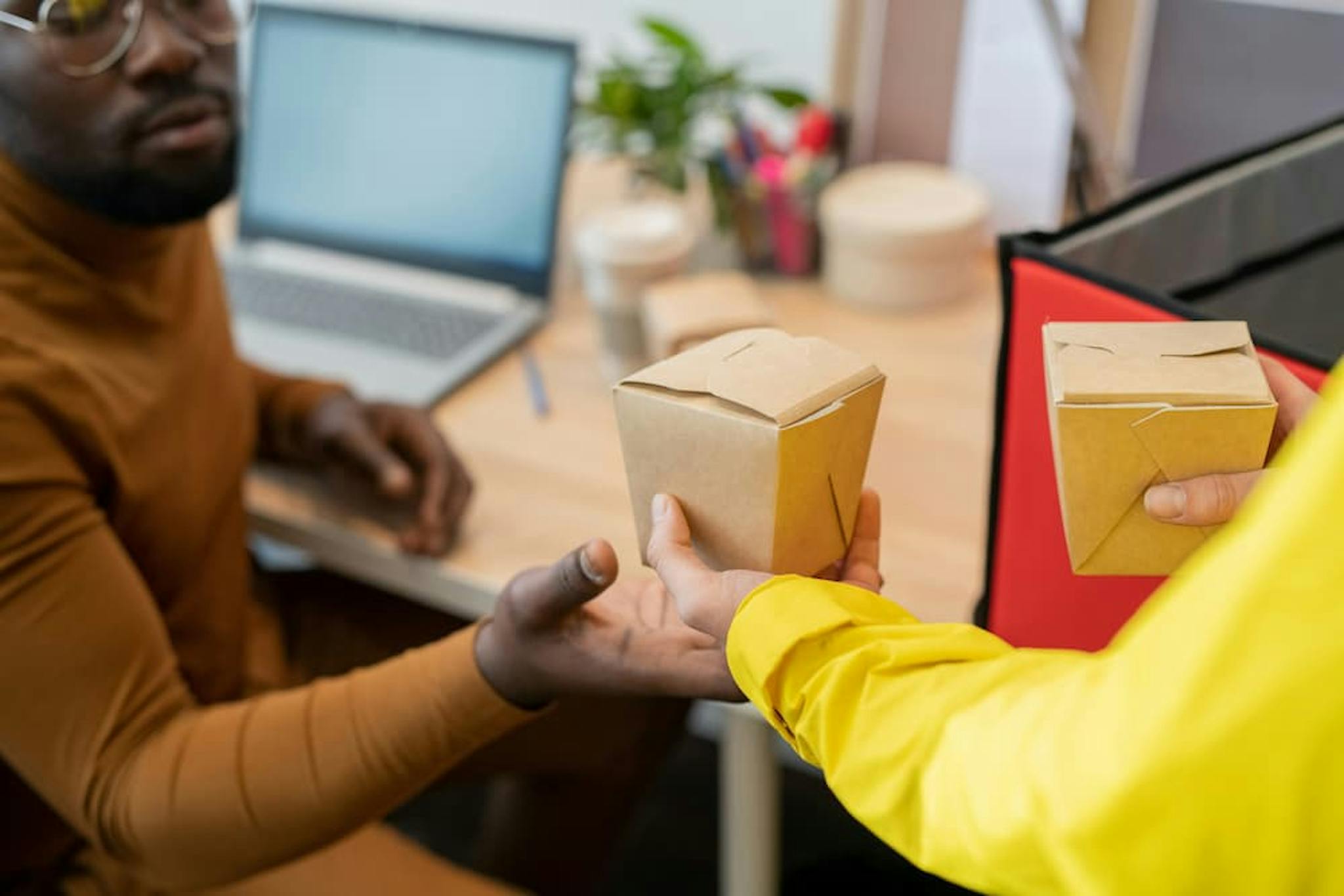 Two people in an office passing food delivery boxes. One person in yellow hands the box to the other, who is wearing a darker color