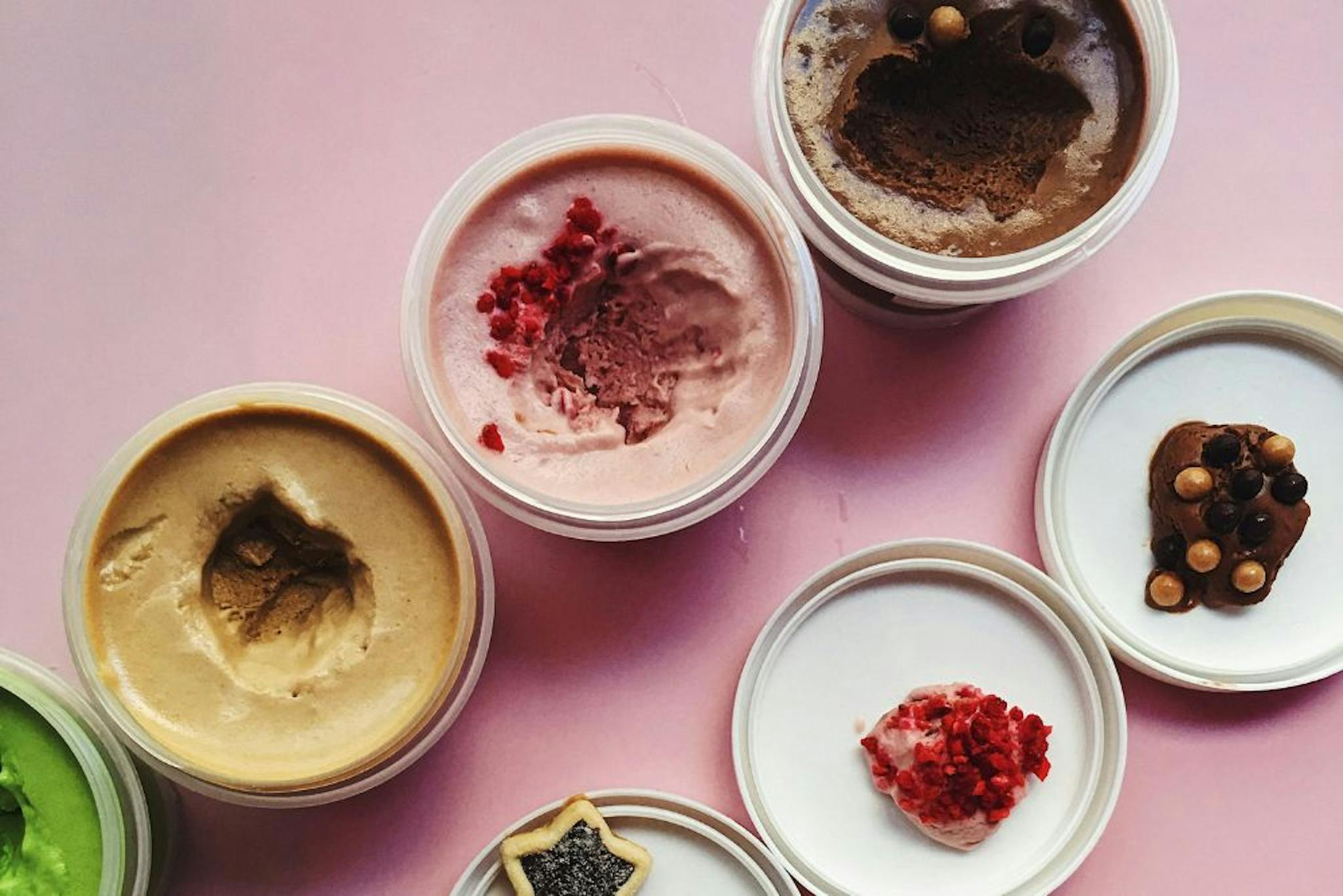 A variety of ice creams in pots, including flavors like chocolate, strawberry, and matcha, arranged colorfully on a pink background